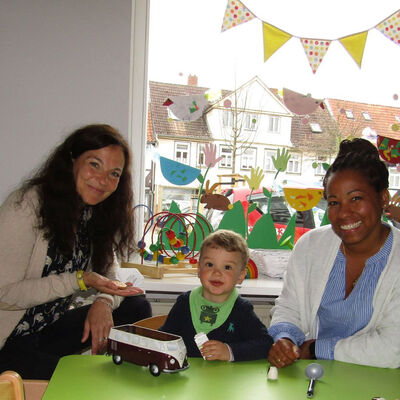 Die Tagespflegemütter Marianela Martinez (r.) und Susann Scholz-Tippner (l.) sitzen mit einem kleinem Jungen am Spieltisch.