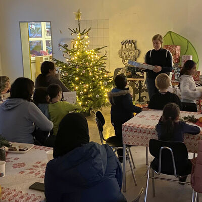 In einem Raum steht ein geschmückter Weihnachtsbaum. Eine Frau hält ein Geschenk in der Hand. An Tischen sitzen mehrere Personen und schauen zu ihr.