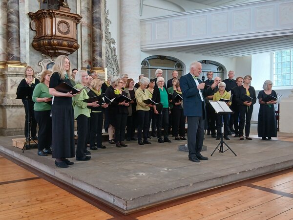 Eine Gruppe von Menschen steht in einer Kirche. Es ist der Chor der Musikschule im Bildungszentrum. Vor der Gruppe steht ein Mann, es ist Chorleiter Kuno Galter. Er gibt sein Abschiedskonzert.