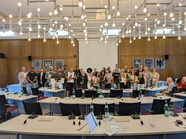 Eine Gruppe von Menschen, Erwachsene und Kinder, stehen in einem Raum. Sie halten Urkunden und Blume. Anlass ist eine Einb�rgerungsfeier,