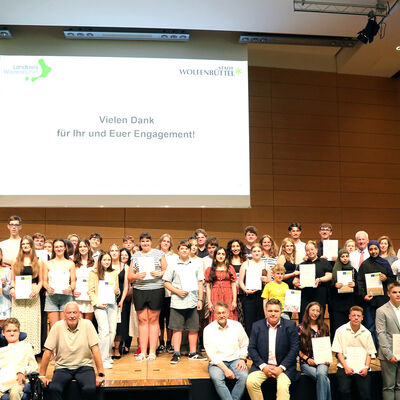 Sch�ler und Sch�lerinnen stehen und sitzen auf einer B�hne. Im Hintergrund stehen auf einem Bildschirm die Worte Vielen Dank f�r Euer Engagement.