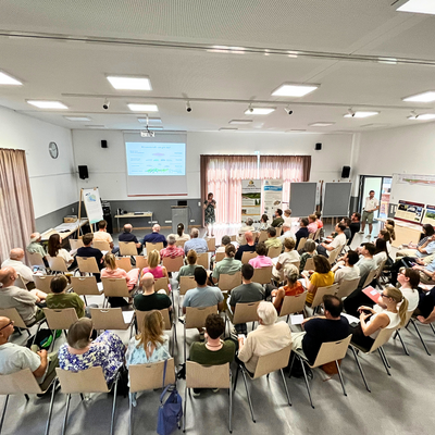 Weitwinkelaufnahme. Menschen sitzen in einem Saal auf St�hlen in mehreren Reihen. Vorne steht eine Person und pr�sentiert ein Thema mit Hilfe einer Leinwand.