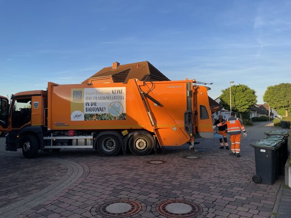 Ein orangefarbener M�llwagen steht auf einer gepflasterten Stra�e. Im Hintergrund blauer Himmel. Zwei M�llwerker sammeln M�lltonnen ein.
