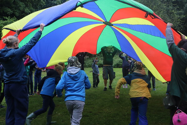 Eine Gruppe von Kindern und Erwachsenen stehen auf einer Wiese im Wald im Kreis, sie halten ein buntes Tuch in den H�nden und schleudern es in die Luft. Einige Kinder laufen darunter hindurch.