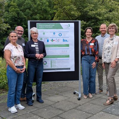 Ein Team hat das Wasserversorgungskonzept gemeinsam erarbeitet: Claudia Ferrara (Untere Wasserbeh�rde, Landkreis Wolfenb�ttel), Umweltdezernent Sven Volkers, Landr�tin Christiana Steinbr�gge, Umweltamtsleiterin Kristina E�, Ferdinand Flechtner (DHI WASY GmbH) und Antje Dreblow-Wulf (Untere Wasserbeh�rde, Landkreis  Wolfenb�ttel). Sie stehen mit einer Tafel, auf der das Konzept visualisiert ist, auf Pflastersteinen vor B�umen.