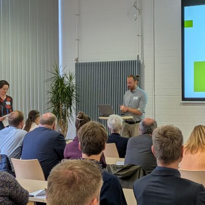Menschen h�ren ein Vortrag zum Wasserversorgungskonzept in einem Saal. Sie sitzen in mehreren Reihen mit dem R�cken zur Kamera. Vorne steht der Vortragende. An der Wand ist eine Pr�sentation zum Thema zu sehen.