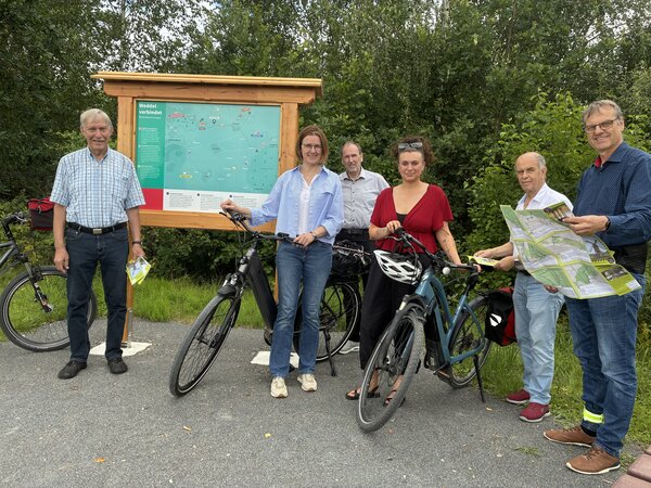 Sechs Personen stehen mit drei Fahrr�dern vor einer Tafel mit Holzrahmen und einer Information auf t�rkisem Grund mit wei�er Schrift. Eine Person h�lt eine Landkarte in der Hand.