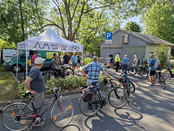B�ume und blauer Himmel im Hintergrund. Ein graues Holzhaus rechts im Bild. Im Vordergrund Fahrradfahrer, die auf einer asphaltierten Fl�che neben ihren R�dern stehen. Sie warten auf den kostenlosen Rad-Check. Ein Zelt mit der Aufschrift Radhaus am Elm steht mittig links auf der Fl�che.