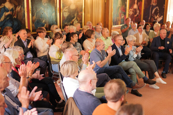 Menschen sitzen in einem Saal, sie klatschen in die H�nde. Im Hintergrund h�ngen verschiedene Gem�lde.