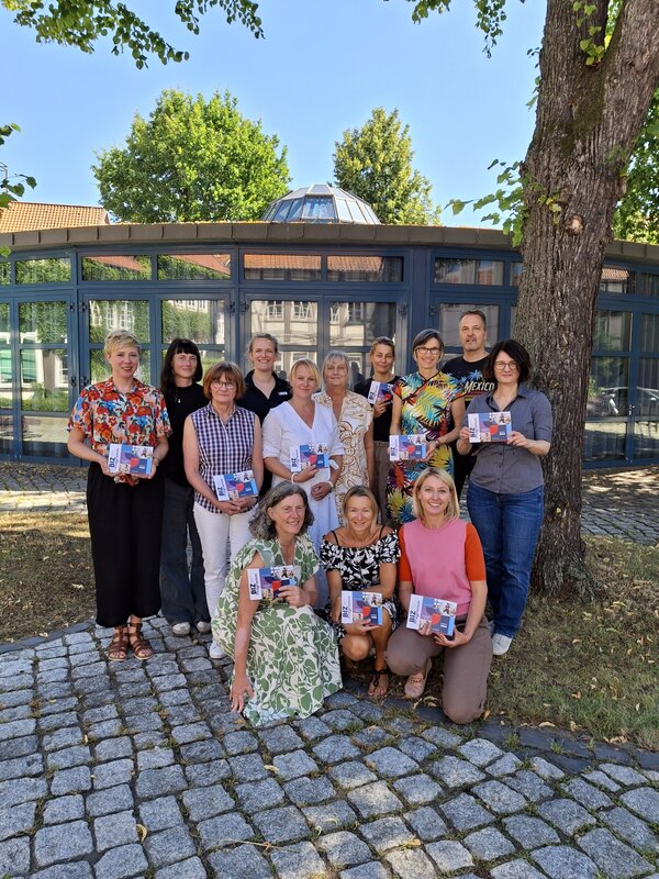 Eine Gruppe von Menschen steht vor einem Geb�ude mit gro�en Fenstern. Ganz vorne knieen drei Menschen. Rechts im BIld ist ein Baum.