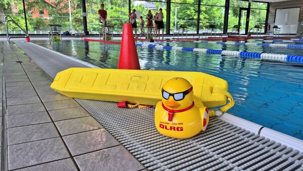 Es ist eine gro�e Gummiente und ein BGer�t f�r die Wasserrettung zu sehen. Darauf der Schriftzug DLRG. Dahinter ein Schwimmbecken in einem Hallenbad.