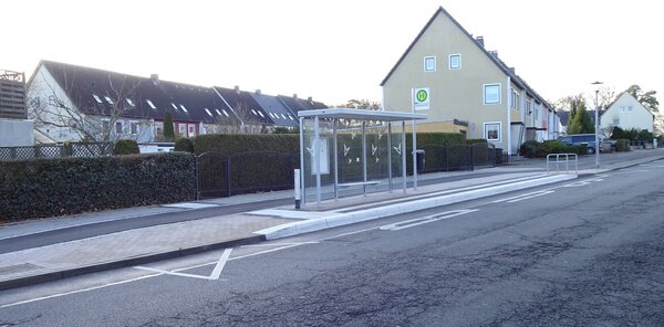 Eine Bushaltestelle, die barrierefrei gestaltet ist. Es ist ein hoher Bordstein in wei� zu sehen. Im Hintergrund stehen H�user.