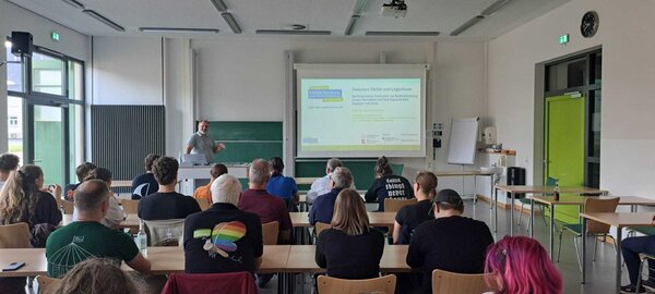 Ein Klassenraum mit Blick zur Leinwand, auf der eine Pr�sentation l�uft. Daneben ein Mann, der sie pr�sentiert. Die Zuschauer sitzen mit dem T�cken zur Kamera und schauen zur Leinwand.