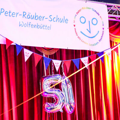 Ein roter Vorhang an einer Stahlkonstruktion. Vor dem Vorhang h&auml;ngt eine Wimpelkette in rot blau und wei&szlig;, daran befestigt ein silberner Luftballon der eine 50 darstellt. Am Stahltr&auml;ger oben h&auml;ngt ein Banner der Peter-R&auml;uber-Schule.