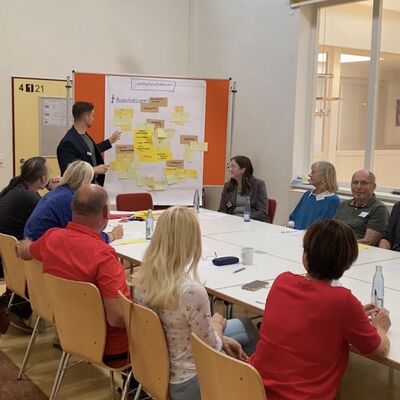 Menschen sitzen an einem langen Tisch und schauen auf eine Tafel. Auf der Tafel h�ngen gelbe und rote beschriebene Zettel.