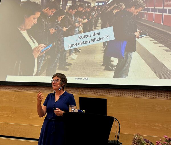 Eine Frau in einem blauen Kleid steht vor einer Leinwand und referiert �ber das Thema "Kultur des gesenkten Blickes". Auf der Leinwand sieht man viele Menschen, die auf ihr Smartphone gucken.