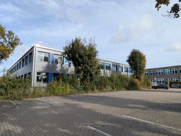 Ein Schulgeb�ude. Davor zwei B�ume. Ganz im Vordergrund ist eine graue Fl�che, das ist ein Parkplatz.