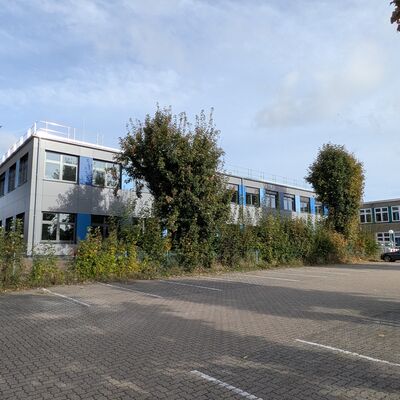 Ein Schulgeb�ude. Davor zwei B�ume. Ganz im Vordergrund ist eine graue Fl�che, das ist ein Parkplatz.