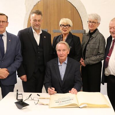 Eine Gruppe von Menschen steht an einem Tisch, eine Person sitzt. Vor dieser Person ist ein gro�es Buch.